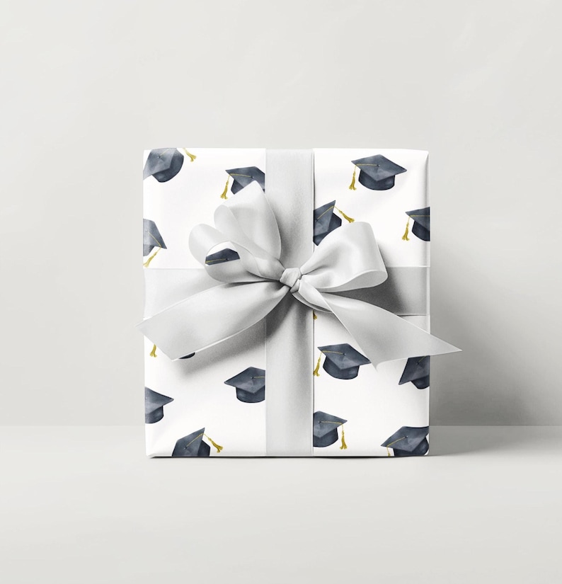 K&ouml;nnte beinhalten: Eine wei&szlig;e Geschenkbox mit einer silbernen Schleife und einem Muster aus schwarzen Abschlusskappen. Die Box ist in wei&szlig;es Papier mit einem sich wiederholenden Muster aus schwarzen Abschlusskappen verpackt.
