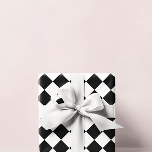 May include: A wrapped gift box with a black and white diamond pattern. The box is tied with a white satin ribbon and bow. The background is a solid light pink.
