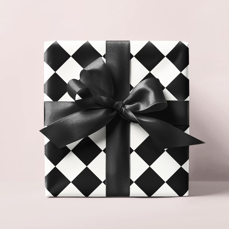 May include: A square gift box wrapped in black and white diamond patterned paper. A wide black satin ribbon is tied around the box, forming a large bow on top. The background is a soft pink.
