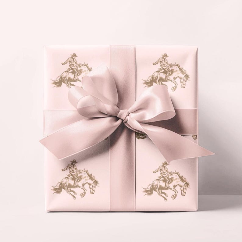 May include: A wrapped gift box with a light pink background and a matching satin ribbon tied in a bow. The wrapping paper features a repeating pattern of a cowboy riding a horse in a gold color.