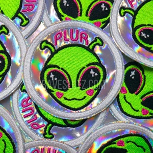 May include: A set of green alien embroidered iron-on patches with pink accents and the word "PLUR" on a silver holographic background.
