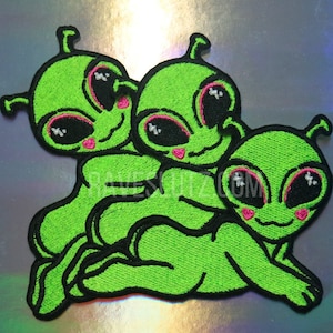 May include: Three green alien embroidered patches with pink hearts and black eyes. The aliens are smiling and have a playful expression.