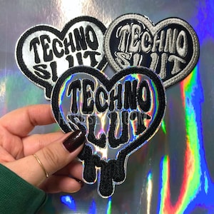 May include: Heart-shaped patches with the words "TECHNO SLUT" in a bold, stylized font. The patches have a black border and the text is in a contrasting color. The background is a holographic, iridescent material.