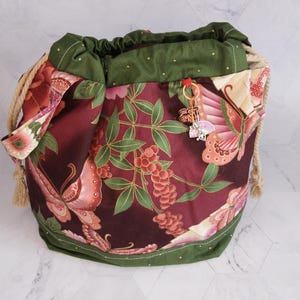 May include: A drawstring bag with a burgundy floral pattern featuring butterflies and foliage. The bag has a green trim and drawstring, with rope handles and a charm. The bag is approximately 25 cm tall.