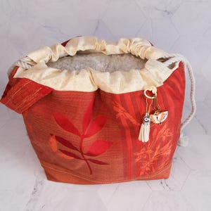 May include: A drawstring bag featuring a red and orange leaf pattern. The bag has a cream-coloured drawstring and trim. A small charm with a butterfly and tassel hangs from the side. The bag is made of fabric.