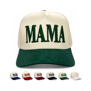 May include: A cream-colored trucker hat with a green brim and the word "MAMA" embroidered in large, dark green letters. The hat has a mesh back and a curved brim. Several other hats with the word "MAMA" are displayed below in different colors.