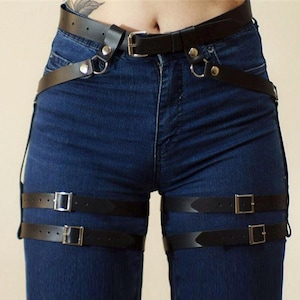 May include: Black leather thigh harness with adjustable straps and buckles. The harness is worn over a pair of blue jeans.