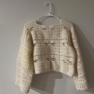 Può includere: Un maglione bianco lavorato a uncinetto con una vestibilità ampia e un motivo quadrato. Il maglione ha una scollatura ampia e maniche lunghe.