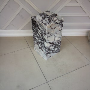 May include: A white marble pedestal with brown and gray veining. The pedestal is square and has a smooth, polished surface.