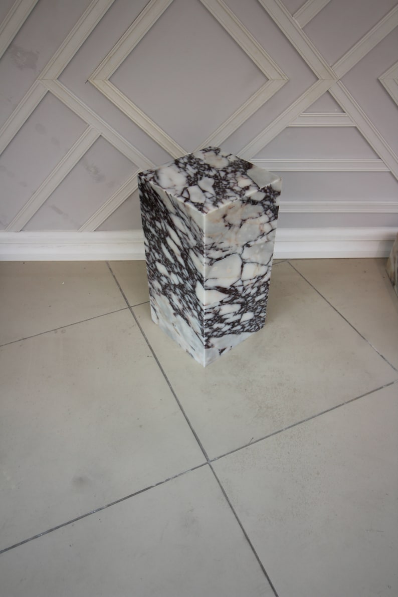 May include: A white marble pedestal with brown and gray veining. The pedestal is square and has a smooth, polished surface.