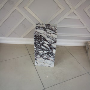 May include: A white marble pedestal with grey and purple veining. The pedestal is square and has a smooth surface.