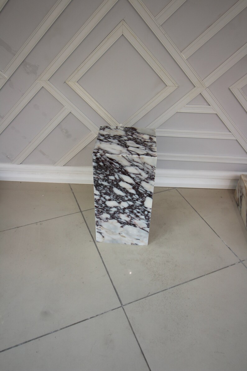 May include: A white marble pedestal with grey and purple veining. The pedestal is square and has a smooth surface.