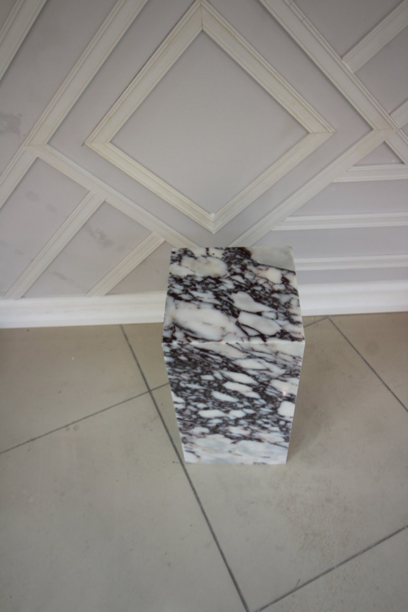 May include: A white marble pedestal with purple and gray veining. The pedestal is square and has a smooth, polished surface.