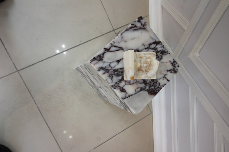 May include: A white marble pedestal with a white and brown marble top. The pedestal has a white marble sculpture on top.