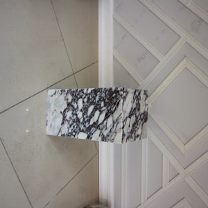 May include: A white marble pedestal with black and gray veining. The pedestal is rectangular and has a smooth, polished surface.