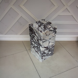 Puede incluir: Un pedestal cuadrado de mármol con un patrón de mármol blanco y morado. El pedestal está sobre un suelo de baldosas de color gris claro.