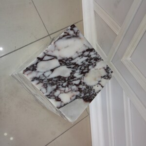 May include: A white marble pedestal with a square top and a swirling pattern of dark red and purple veins.