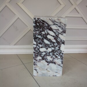 May include: A white marble pedestal with a pink and grey marbling pattern. The pedestal is square and has a smooth surface.