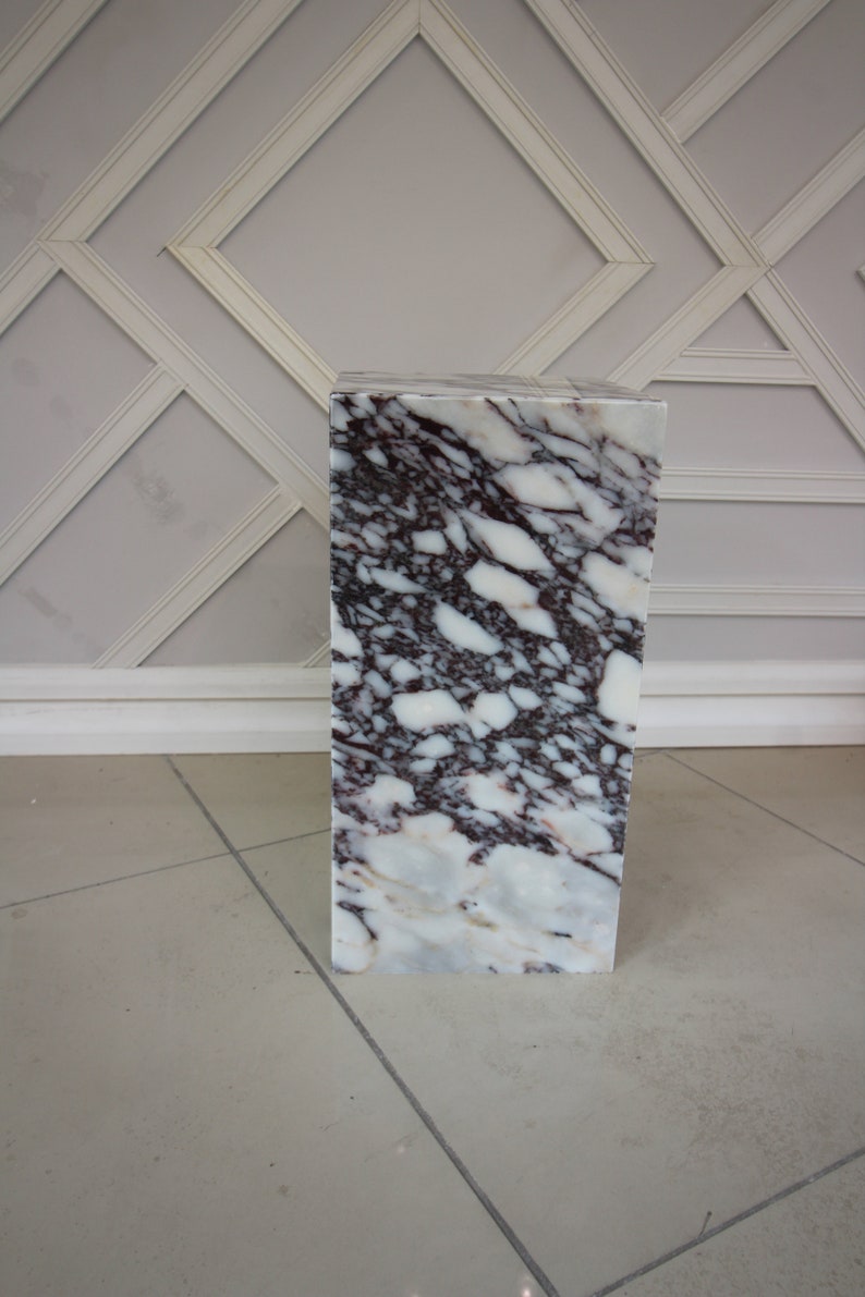 May include: A white marble pedestal with a pink and grey marbling pattern. The pedestal is square and has a smooth surface.