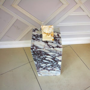 May include: A white marble pedestal with a pink and white marbled pattern. A small white sculpture of a head sits on top of the pedestal.