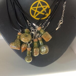 May include: A collection of glass potion bottle necklaces with cork stoppers, displayed on a black mannequin. The bottles are various shapes and sizes, filled with amber and green liquids. A yellow pentagram is painted on the mannequin.