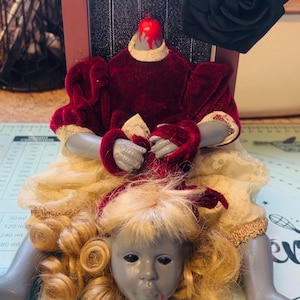 Könnte beinhalten: Ein puppenartiges Kunstwerk mit einem kopflosen Torso in einem bordeauxfarbenen Samtkleid mit Spitzenbesatz. Der Puppenkopf mit blonden Locken ruht unterhalb des Torsos. Eine schwarze Rose und ein bordeauxfarbener Rahmen vervollständigen die makabre Komposition.