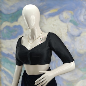 May include: A black, sleeveless blouse with a V-neckline and a button closure. The blouse has a short, fitted style and is made of a smooth, silky fabric. The blouse is displayed on a mannequin with a gold chain necklace.
