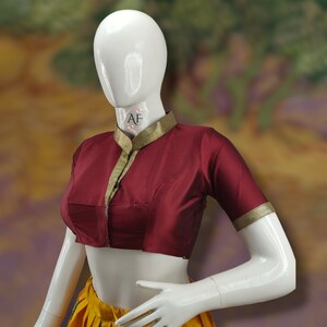 May include: A maroon blouse with a high, mandarin collar and short sleeves. The collar and sleeves are trimmed with gold fabric. The blouse is on a white mannequin.