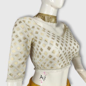 May include: White blouse with gold floral pattern and elbow-length sleeves. The blouse has a V-neck and is trimmed with silver and white fringe. A gold choker necklace with fringe is also visible.