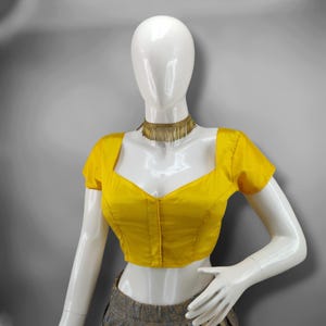 May include: A bright yellow blouse with short sleeves and a square neckline is displayed on a mannequin. The blouse has a fitted silhouette and a button-up front. A gold-colored necklace with fringe detail adorns the mannequin's neck. The mannequin is wearing a gray and gold patterned skirt.