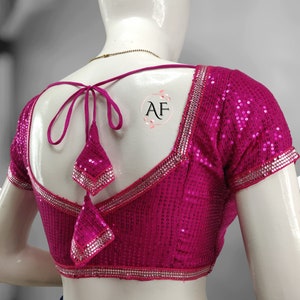 Puede incluir: Una blusa rosa con lentejuelas con escote en V y mangas cortas. La blusa tiene un ribete decorativo de lentejuelas plateadas y un lazo en la espalda.