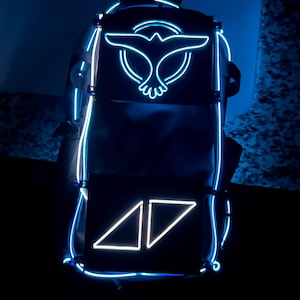 Puede incluir: Mochila negra con contorno y detalles en azul brillante. La mochila presenta un logotipo de pájaro y las letras "AV" en neón blanco. Tiene un asa superior y correas ajustables.