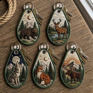 May include: Embroidered teardrop-shaped keychains featuring forest scenes with wildlife. Designs include a bear, deer, wolf, fox, and moose, each set against a backdrop of trees, mountains, and celestial elements. Keychains are attached to metal key rings.