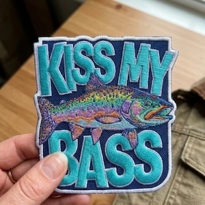 May include: Embroidered patch with the text "KISS MY BASS" in turquoise letters on a navy blue background. A colorful fish illustration is centered within the patch. The patch is held by a hand, with a tan jacket in the background.