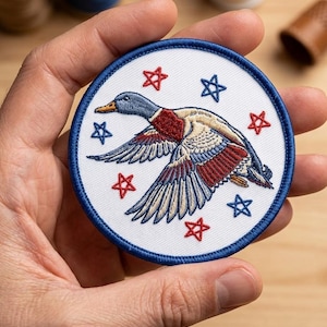 May include: Round embroidered patch with a blue border, featuring a duck in flight. The design incorporates red, white, and blue colours, with stars. The patch has a patriotic theme.