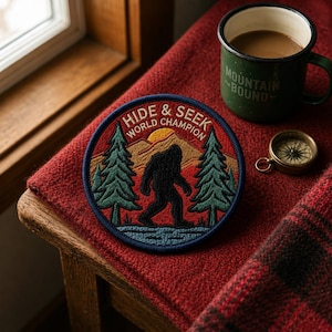 May include: A round embroidered patch with the text "HIDE & SEEK WORLD CHAMPION" and a silhouette of Bigfoot. The patch is on a red blanket next to a green mug with the words "MOUNTAIN BOUND" and a small compass.