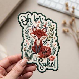 May include: Embroidered patch with a red fox and floral design, with the text "Oh for Sake!". The patch has a green scalloped border. The fox is surrounded by leaves and flowers.