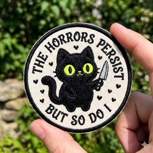 May include: Circular patch featuring a black cat with green eyes holding a knife, surrounded by the text "THE HORRORS PERSIST BUT SO DO I." The patch has a cream background with black text and heart accents.