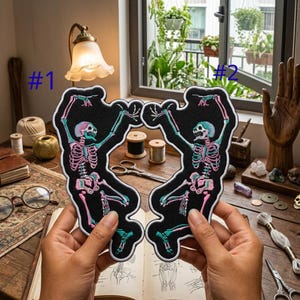 May include: Two black embroidered patches featuring a dancing skeleton design. The skeletons have pink and teal accents, with a white border. The patches are held up in front of a wooden table with other craft supplies.