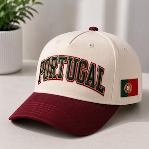 May include: A cream-coloured baseball cap with a dark red brim and a dark red button on top. The front features the word PORTUGAL embroidered in dark green with a dark red outline. A small rectangular flag of Portugal is on the side panel.