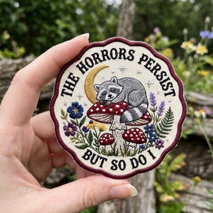 May include: Embroidered patch with a raccoon sleeping on a red mushroom with white spots. The patch has a cream-colored background with the text "THE HORRORS PERSIST BUT SO DO I" in a circular design, surrounded by flowers and a crescent moon.