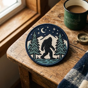 May include: A circular navy blue patch with a black Bigfoot silhouette between two green pine trees. The design includes a crescent moon, stars, and a mountain range. A green enamel mug with "MOUNTAIN BOUND" is visible.