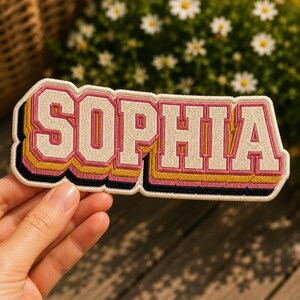 May include: Embroidered patch in a retro style, featuring the name "SOPHIA" in a bold, layered design. The letters are white with pink and yellow outlines, and a black border. This patch is likely for clothing or accessories.