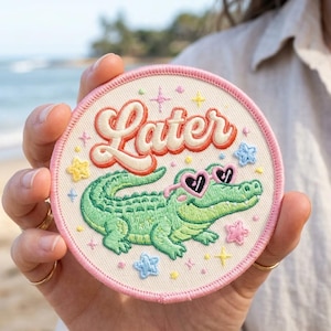 May include: Embroidered patch with a light pink border and the word "Later" in a retro font. The patch features a green alligator wearing heart-shaped sunglasses. Decorative stars and flowers surround the alligator.