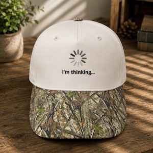 May include: A baseball cap with a white front panel and a camouflage patterned brim. The front panel features an embroidered grey circular loading icon above the black text that reads, I'm thinking... The cap has a button on the top center.