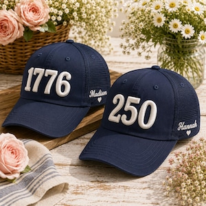 May include: Two navy blue baseball caps with mesh back panels. One cap features the white embroidered text 1776 and the name Madison with a heart. The other cap features the white embroidered text 250 and the name Hannah with a heart.