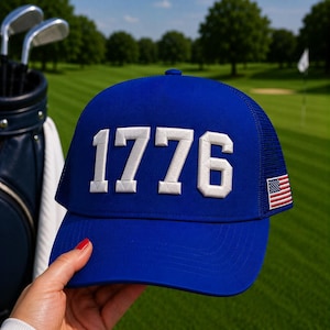 以下が含まれることがあります： 白で刺繍された数字「1776」が入った青いトラッカーハット。帽子はメッシュの背面と小さなアメリカ国旗のパッチが付いています。背景にはゴルフクラブとゴルフバッグが見えます。