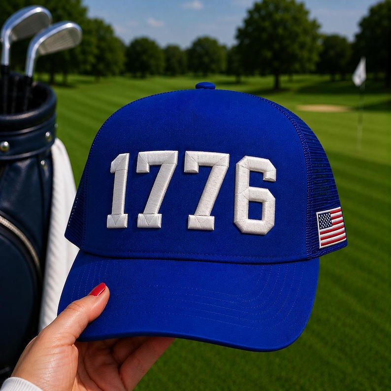 以下が含まれることがあります： 白で刺繍された数字「1776」が入った青いトラッカーハット。帽子はメッシュの背面と小さなアメリカ国旗のパッチが付いています。背景にはゴルフクラブとゴルフバッグが見えます。