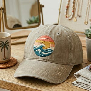 May include: A khaki baseball cap with a circular embroidered patch featuring a sunset over ocean waves. The patch includes shades of yellow, orange, pink, and blue. The cap is on a wooden surface.
