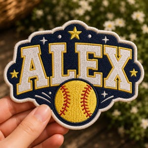 May include: Embroidered patch featuring the name "ALEX" in white, outlined in yellow, on a navy blue background. A yellow softball with red stitching is centered below the name. The patch has a decorative border with stars and crosses.